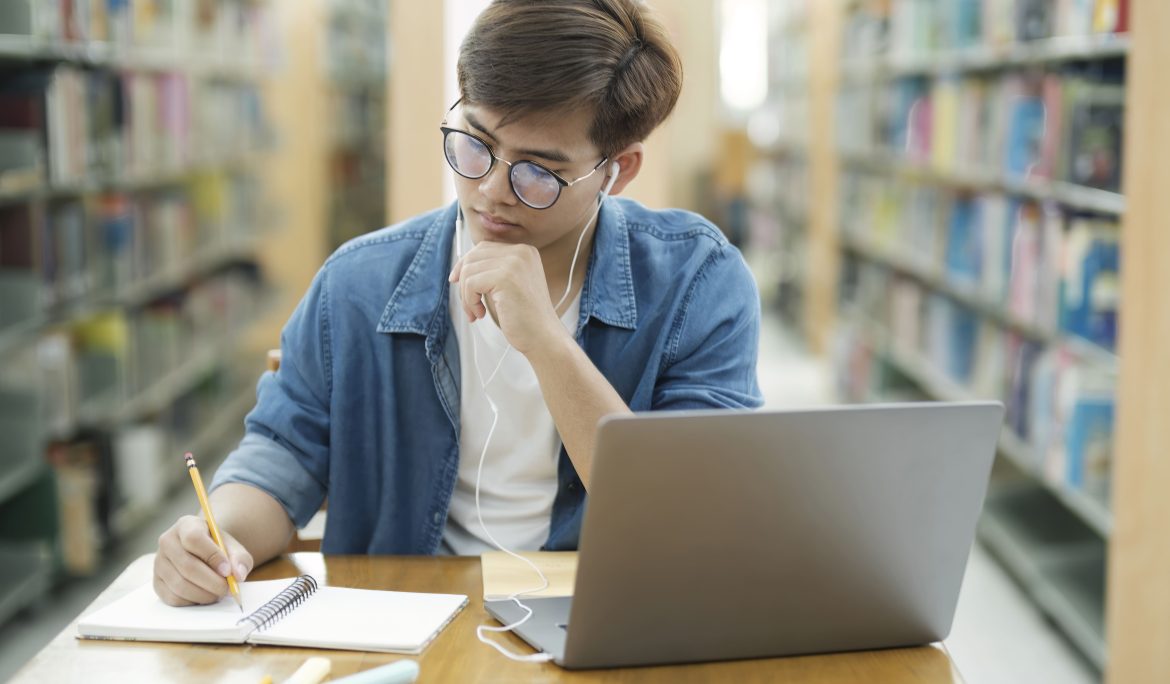 Student taking notes beside a laptop while studying in a library