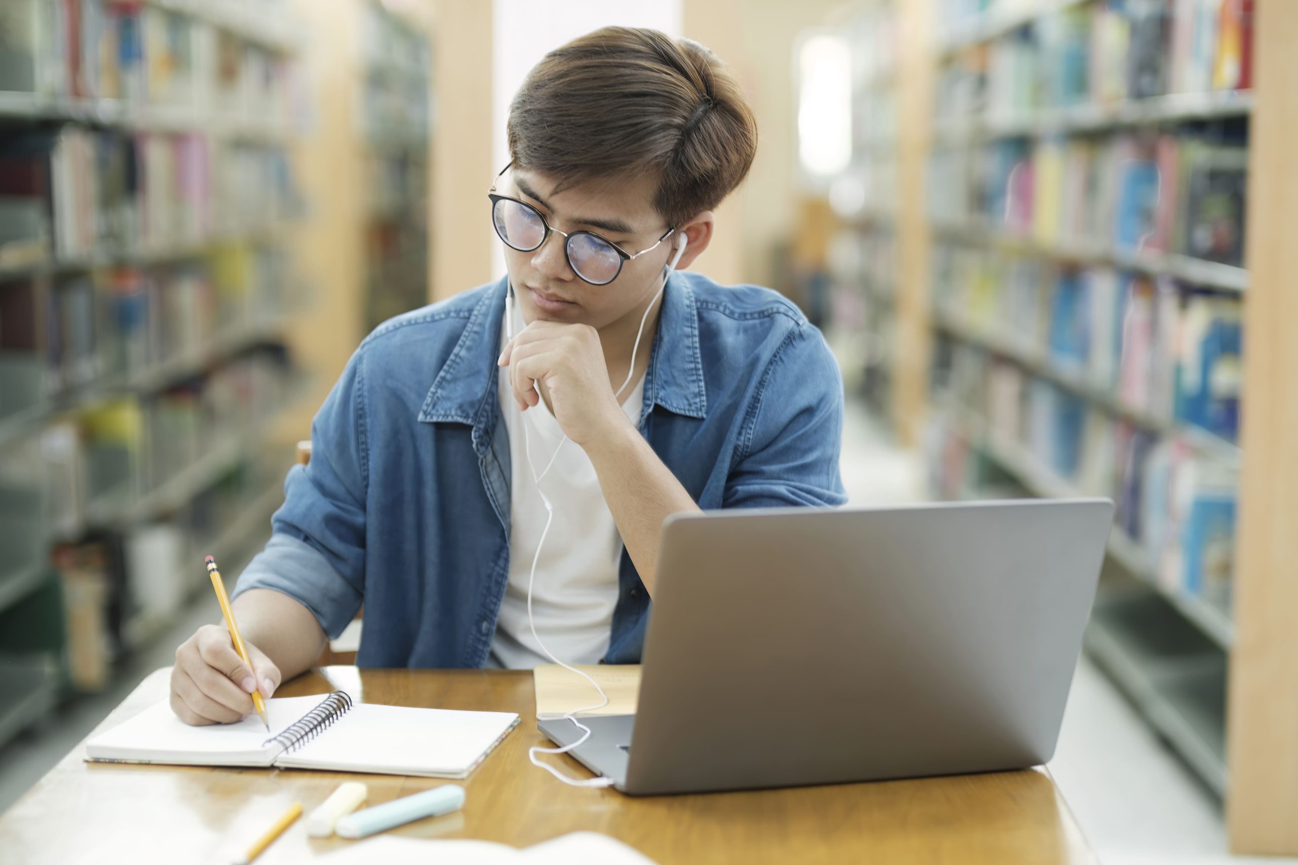 Student taking notes beside a laptop while studying in a library